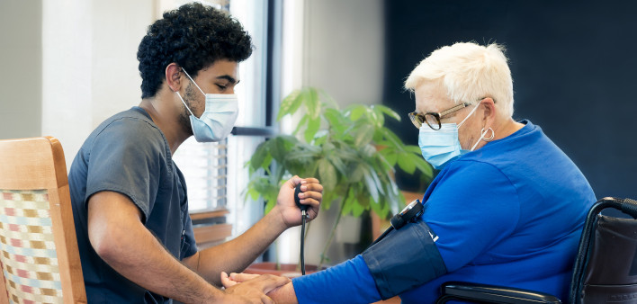 Portrait of a smling African-American male nursing employee wearing gray scrubs and face mask interacts with a senior aged patient during the Covid-19 pandemic,