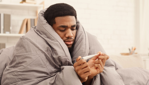 Ill african-american man covered with blanket holding thermometer, having fever