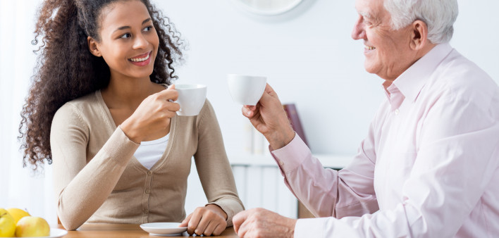 Young happy caregiver and senior smiling man drinking cup of coffee together at home