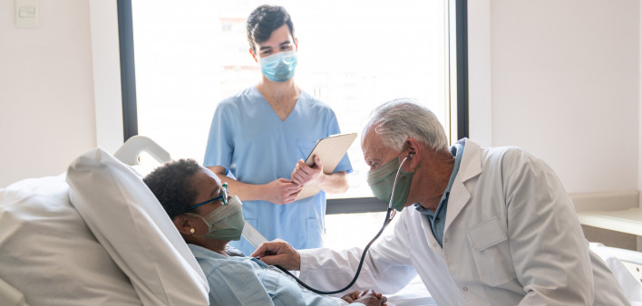 Senior male doctor checking black hospitalized female patient listening to her heart and nurse standing next to them all wearing protective facemasks