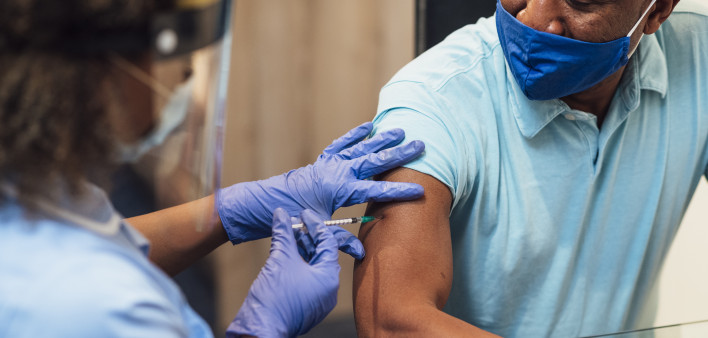 A mixed race female nurse wearing a protective face shield, surgical mask and protective gloves administering the COVID-19 vaccine to a senior black man in his home.