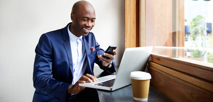 Portrait of happy african businessman using phone while working on laptop in a restaurant