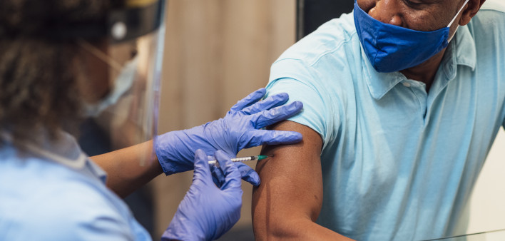 A mixed race female nurse wearing a protective face shield, surgical mask and protective gloves administering the COVID-19 vaccine to a senior black man in his home.