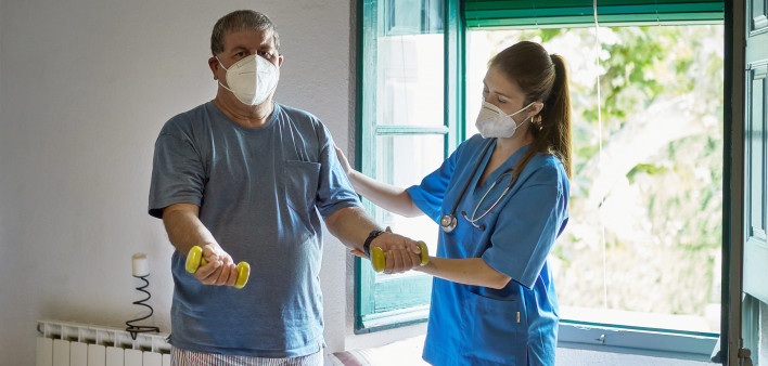 Young healthcare worker in blue scrubs and man in mid 60s wearing protective masks as he lifts lightweight dumbbells during recovery program