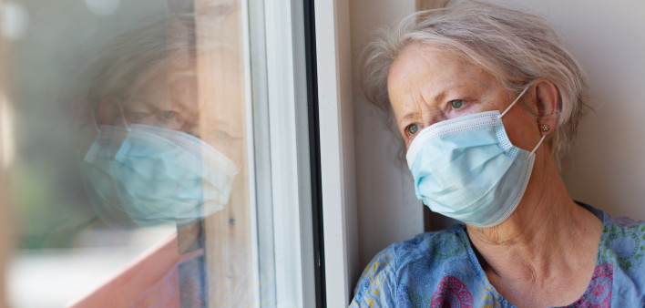 Senior woman with grey hair, wearing a facial protective mask and a blue dress with a pattern, looking out of window during time of self isolation, she is leaning her head against window frame