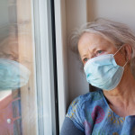 Senior woman with grey hair, wearing a facial protective mask and a blue dress with a pattern, looking out of window during time of self isolation, she is leaning her head against window frame