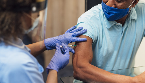 A mixed race female nurse wearing a protective face shield, surgical mask and protective gloves administering the COVID-19 vaccine to a senior black man in his home.