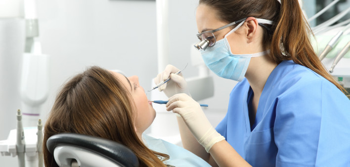 Dentist wearing eyeglasses gloves and mask examining a patient teeth with a dental probe and a mirror in a clinic box with equipment in the background