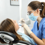 Dentist wearing eyeglasses gloves and mask examining a patient teeth with a dental probe and a mirror in a clinic box with equipment in the background
