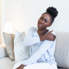 African woman sitting on couch take break touches her shoulder suffers from painful feelings ache caused by poor wrong posture, sedentary work, sitting at laptop for long period