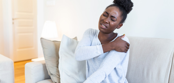 African woman sitting on couch take break touches her shoulder suffers from painful feelings ache caused by poor wrong posture, sedentary work, sitting at laptop for long period