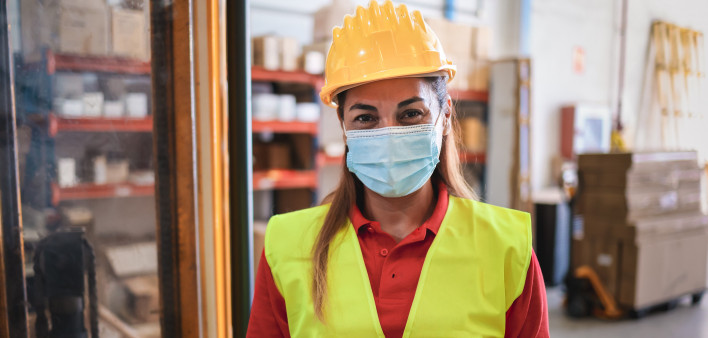Portrait of worker woman holding cardboard box inside warehouse while wearing safety mask