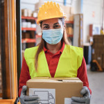 Portrait of worker woman holding cardboard box inside warehouse while wearing safety mask