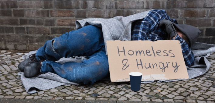 Male Beggar Lying On Street With Homeless And Hungry Text On Cardboard