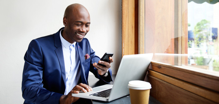 Portrait of happy african businessman using phone while working on laptop in a restaurant