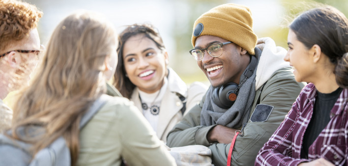 Five young adults are sitting at a picnic table outside on a fall day. They are smiling and chatting