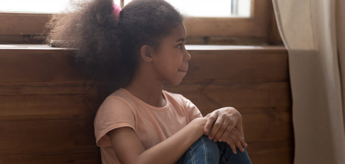 Pensive african preschool or school age girl sitting on floor alone, leaned on wooden wall puts hands on laps looking away