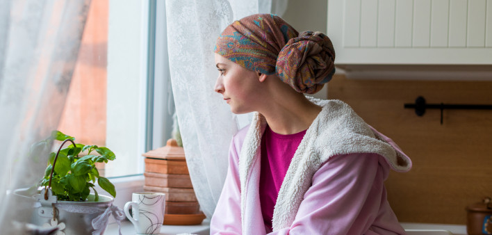 Young adult female cancer patient wearing headscarf and bathrobe sitting in the kitchen with her pet cat, looking out window.