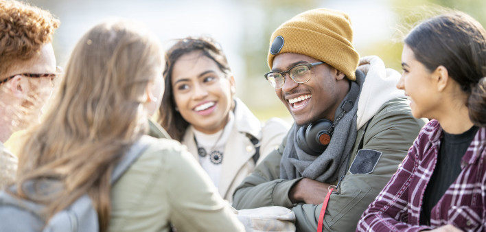 Five young adults are sitting at a picnic table outside on a fall day. They are smiling and chatting