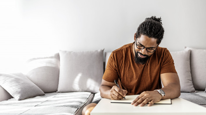 Image of happy African man with notepad and pen sitting on sofa.