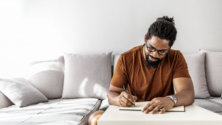 Image of happy African man with notepad and pen sitting on sofa.