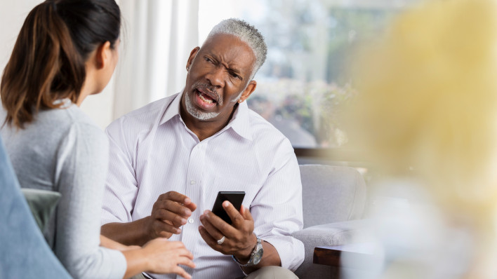 Frustrated senior man holds smartphone while discussing an issue with a female therapist.