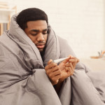 Ill african-american man covered with blanket holding thermometer, having fever
