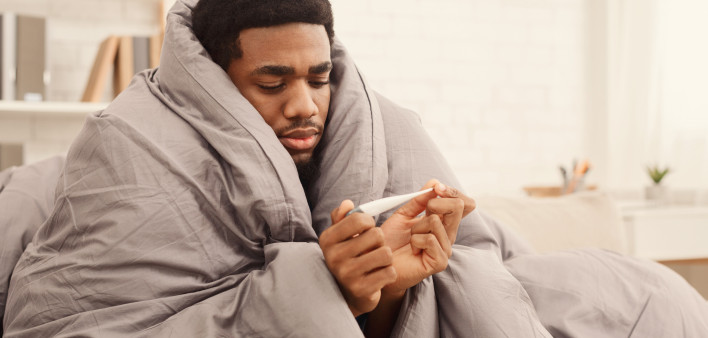Ill african-american man covered with blanket holding thermometer, having fever