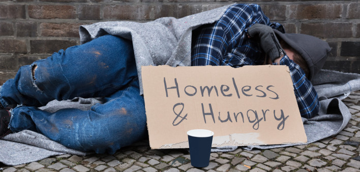 Male Beggar Lying On Street With Homeless And Hungry Text On Cardboard