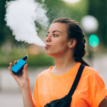 Pretty young hipster woman vape ecig, vaping device at the sunset.