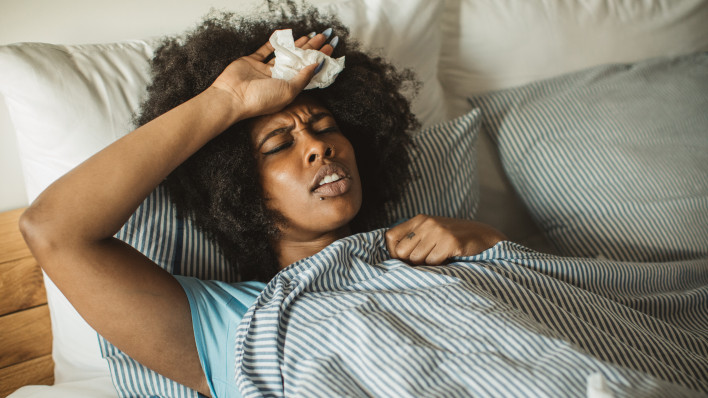 Young African-American woman feeling sick at home. Lying in bed, drinking tea and medicine. Daytime.