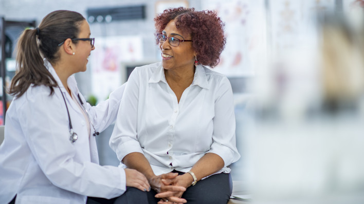 An African American woman smiles lightly as she discusses with the doctor sitting beside her. They are in a medical office.