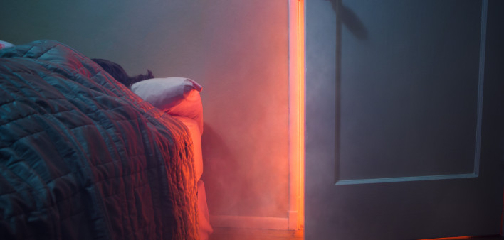 A depiction of a house fire, indoors. Smoke and light from a fire pour through a cracked room door, a person visible sleeping in their bed. Horizontal with copy space.