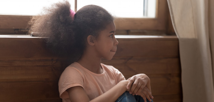 Pensive african preschool or school age girl sitting on floor alone, leaned on wooden wall puts hands on laps looking away