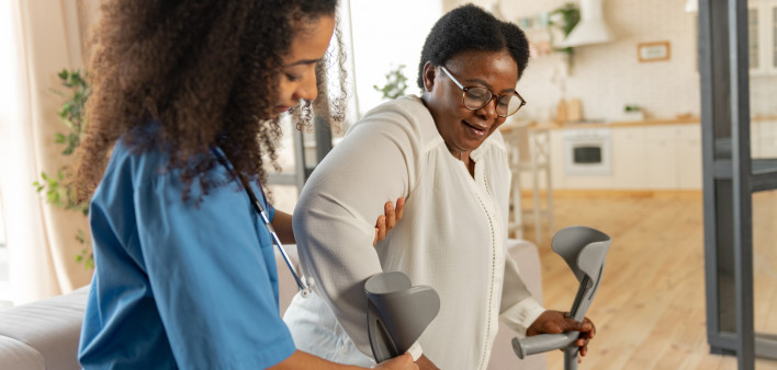 Giving crutches. Curly young dark-haired nurse giving crutches to aged woman wearing glasses after leg surgery