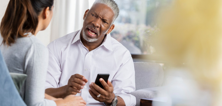 Frustrated senior man holds smartphone while discussing an issue with a female therapist.