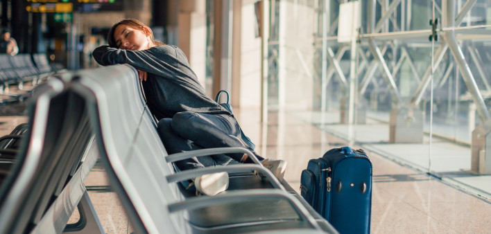 Young tired woman sleeping alone in empty airport with her hand luggage, waiting flight - transportation, low cost traveling, delayed or cancelled flight