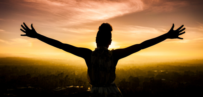 A young African American Woman raises her arms facing the sunset down over the valley and the ocean.