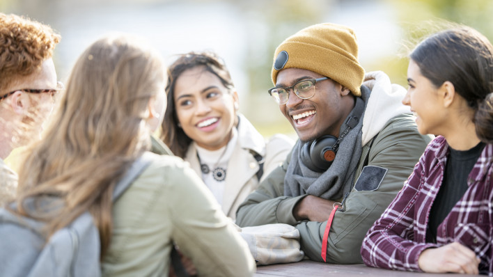 Five young adults are sitting at a picnic table outside on a fall day. They are smiling and chatting