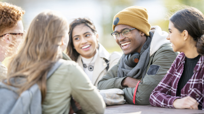 Five young adults are sitting at a picnic table outside on a fall day. They are smiling and chatting