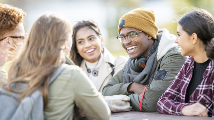Five young adults are sitting at a picnic table outside on a fall day. They are smiling and chatting