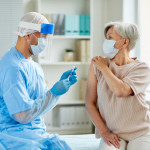 Side view shot of male nurse wearing protective mask and gloves preparing medical syringe for giving injection to senior patient