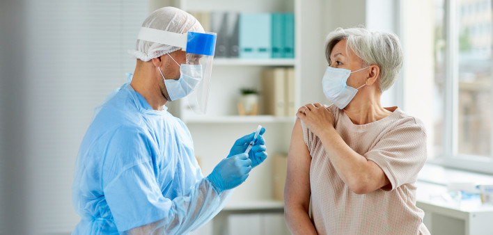 Side view shot of male nurse wearing protective mask and gloves preparing medical syringe for giving injection to senior patient