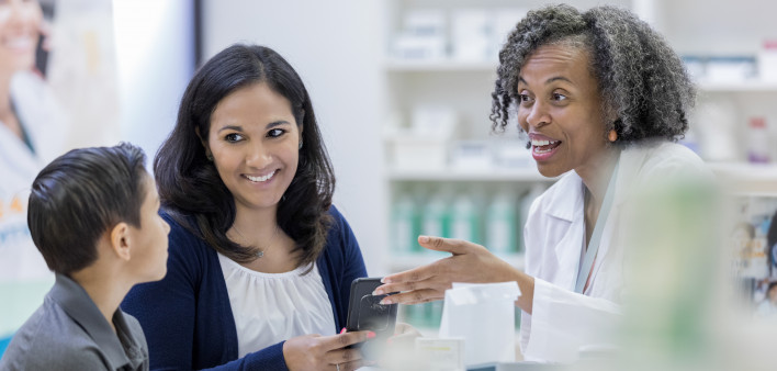 Smiling pharmacist talks withe young boy and his mom about his medication. The boy's mom is holding a smartphone.