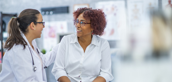 An African American woman smiles lightly as she discusses with the doctor sitting beside her. They are in a medical office.