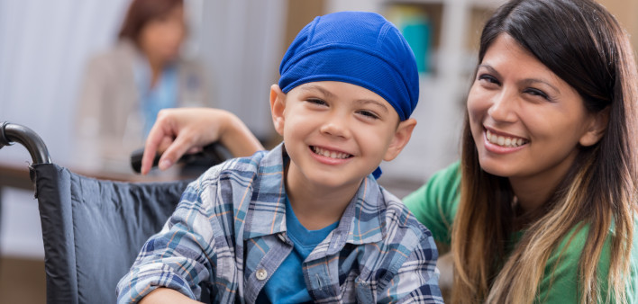 Mid adult mom and her elementary age son wait in waiting room in doctor's office or hospital. The boy is wearing a blue cap and is sitting in a wheelchair.