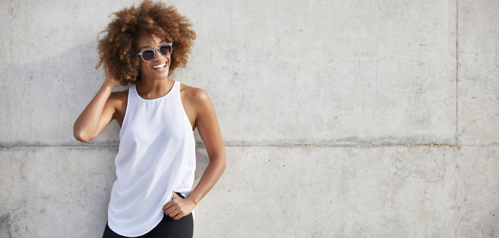Portrait of happy young woman wearing tank top and sunglasses. Beautiful female is with frizzy short hair. She is leaning on wall.