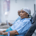 A woman wearing a head scarf lies on a hospital bed and looks to the side in contemplation.