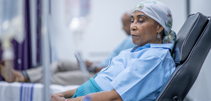 A woman wearing a head scarf lies on a hospital bed and looks to the side in contemplation.