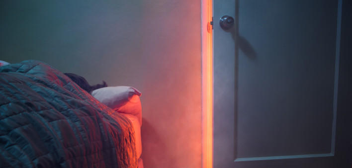 A depiction of a house fire, indoors. Smoke and light from a fire pour through a cracked room door, a person visible sleeping in their bed. Horizontal with copy space.